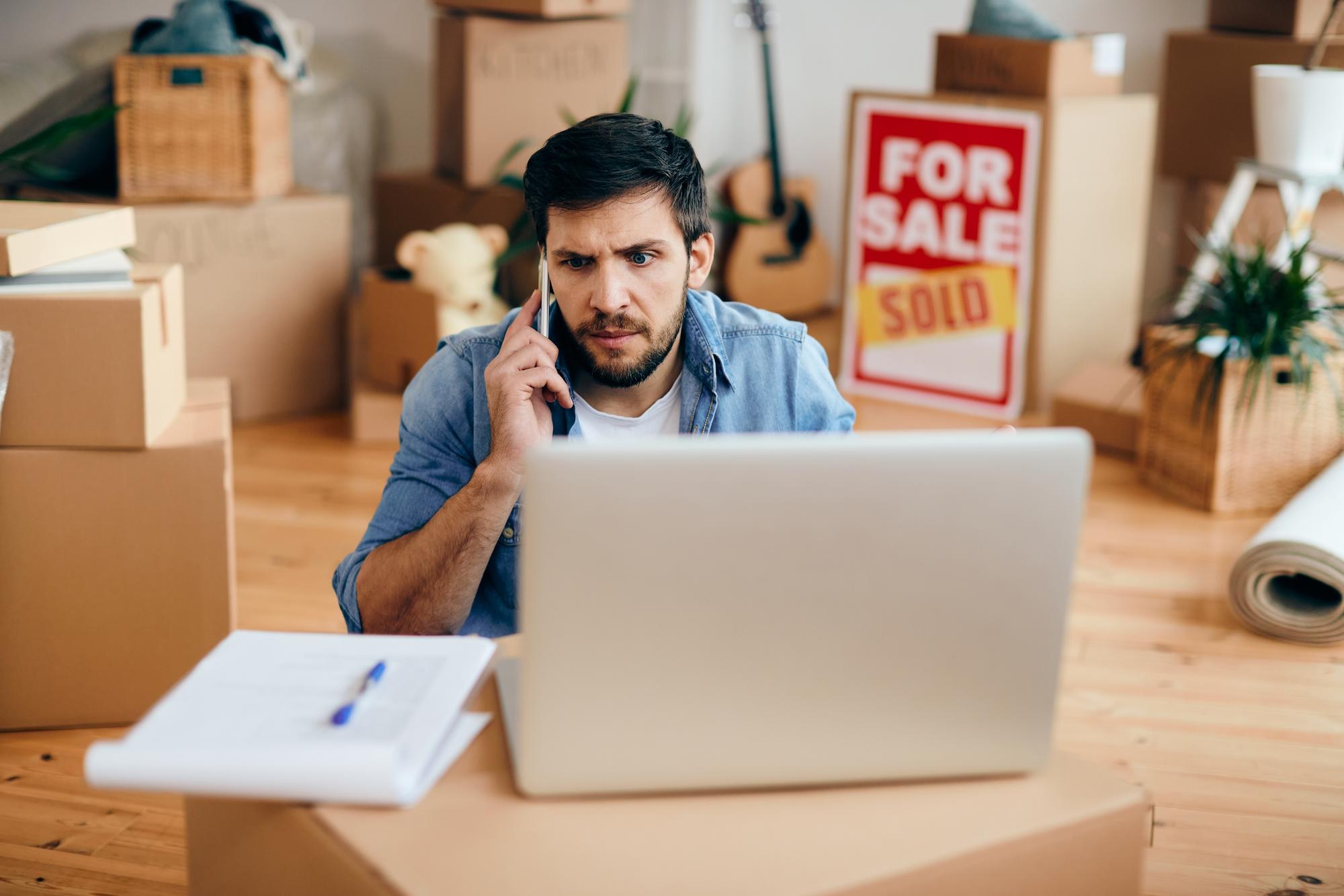 Stressed man speaking on the phone near a property for-sale sign